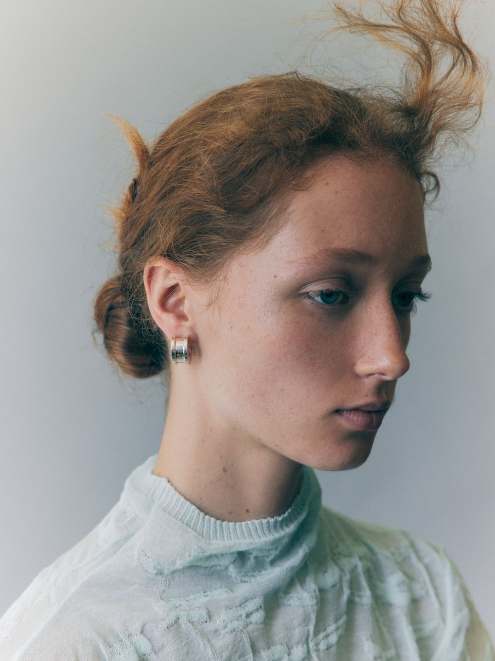 Close-up studio portrait of a young woman with red hair styled in a bun and loose strands, wearing an earring and textured top.