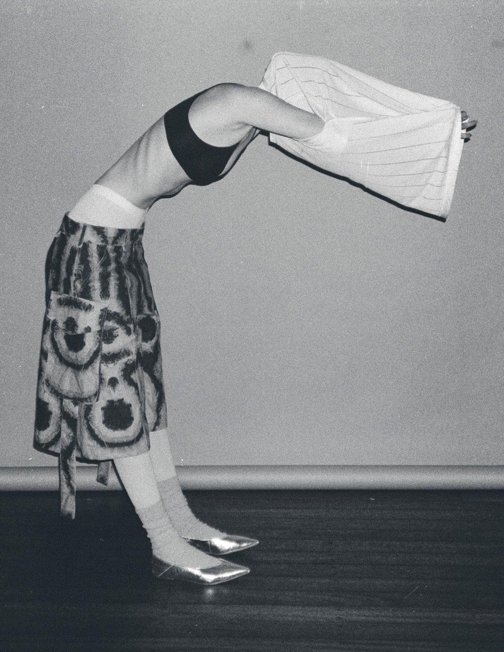An adult female with an arched back wears a bra top, printed skirt, socks, and metallic shoes while holding a striped shirt indoors.