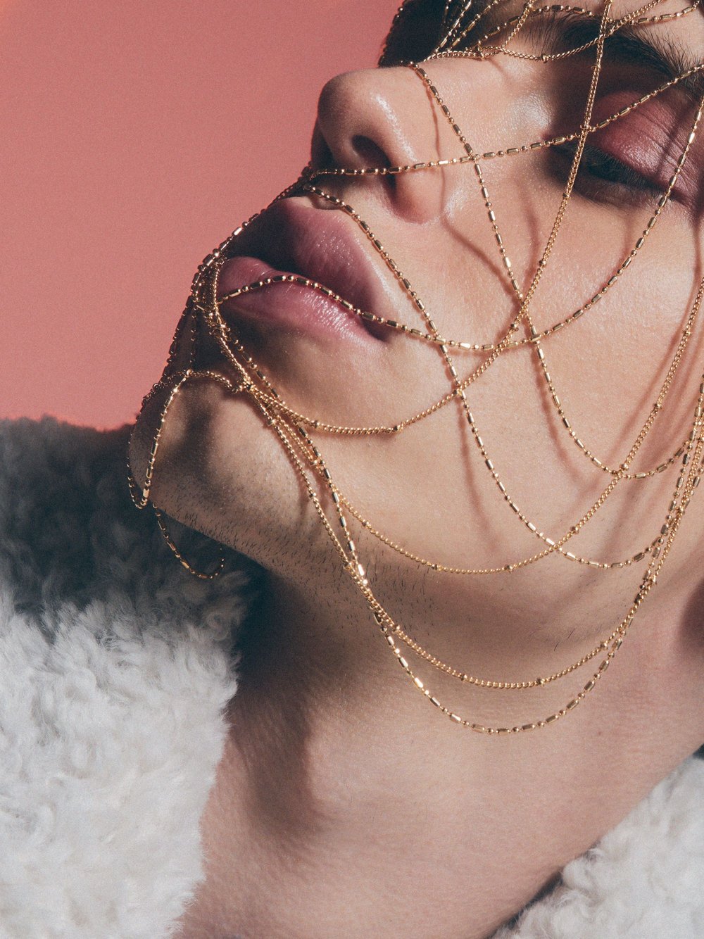 Close-up of a person with layered gold chains draped across their face against a peach background, illuminated by soft studio lighting.