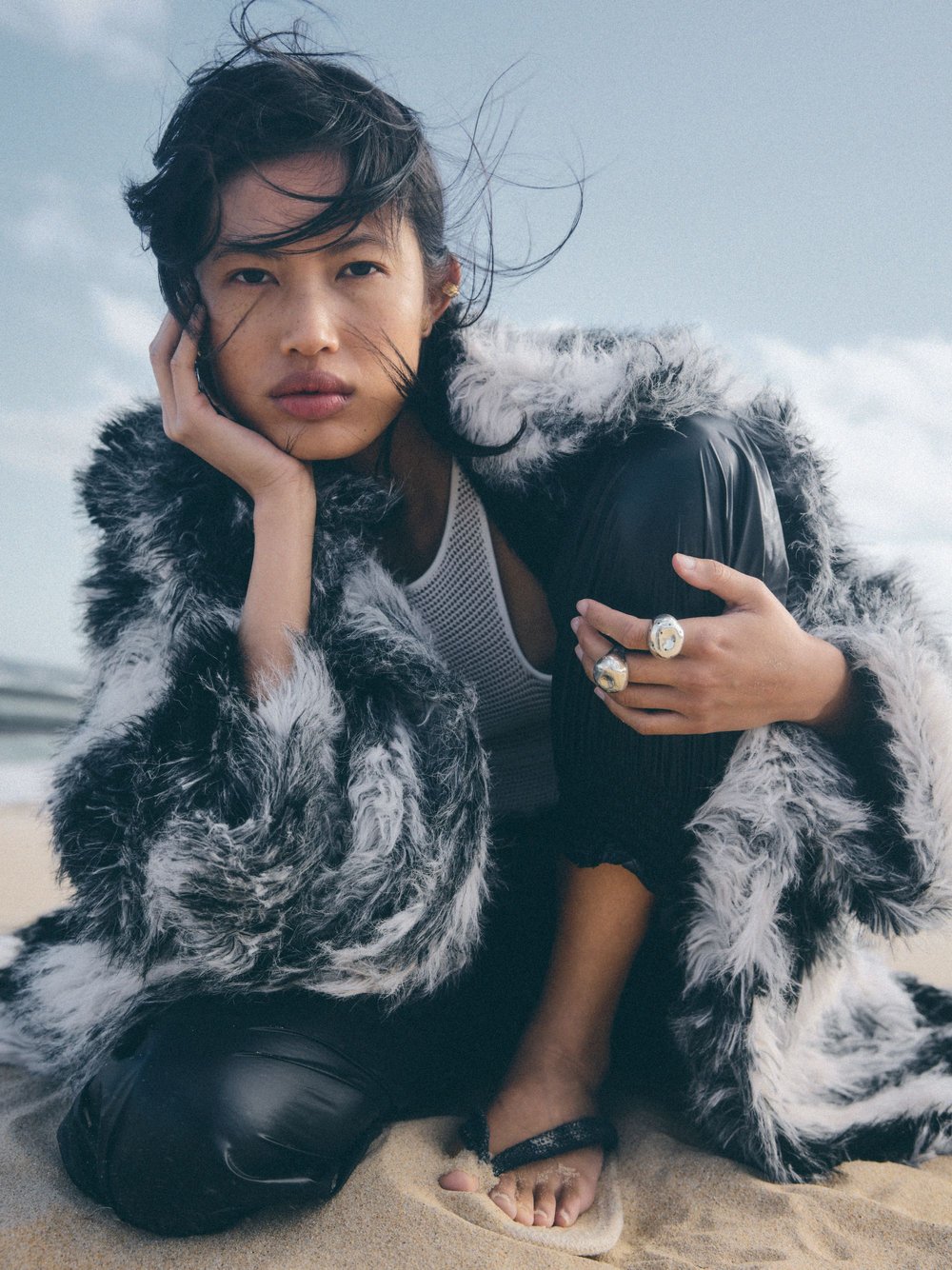 Asian woman poses on sandy beach with a fur-trimmed jacket, rings, and thong sandals against a blue sky and ocean background.