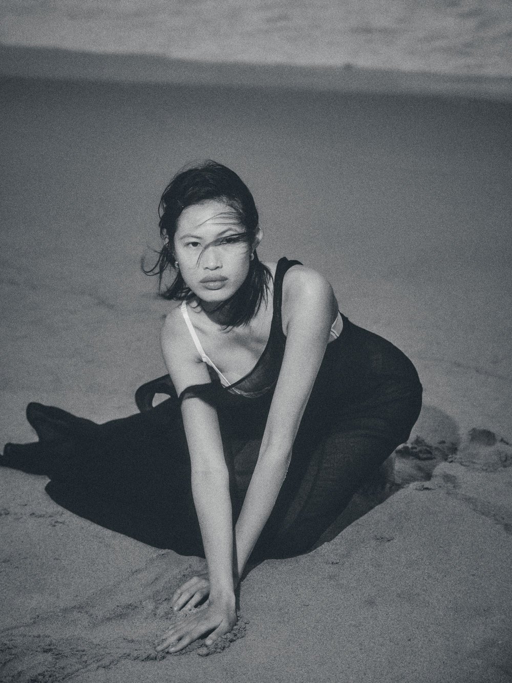 Monochromatic portrait of a young Asian woman in a dark dress kneeling on a sandy beach with soft natural lighting and a calm sea.