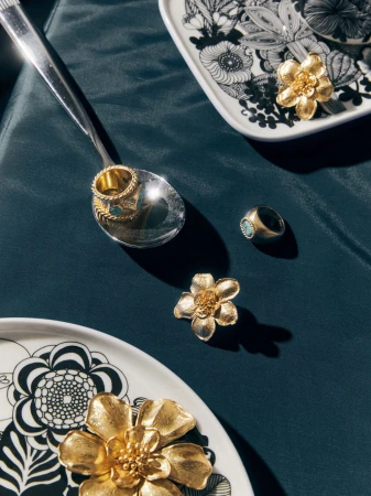 Close-up of gold rings on a silver spoon and gold flower ring holders displayed on a dark blue tablecloth with patterned plates.