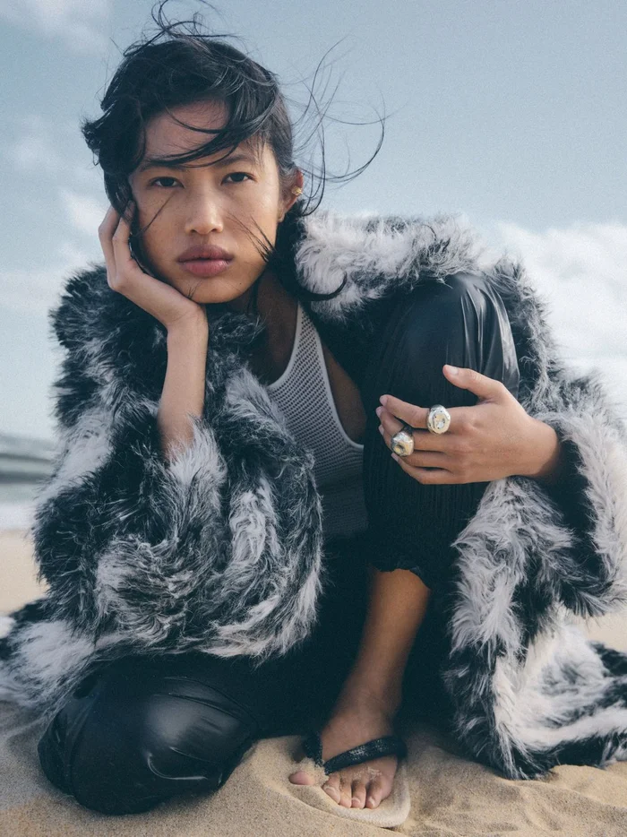 Woman in a black and white faux fur coat and ribbed tank top crouching on a sandy beach, photographed by Seung Rok.
