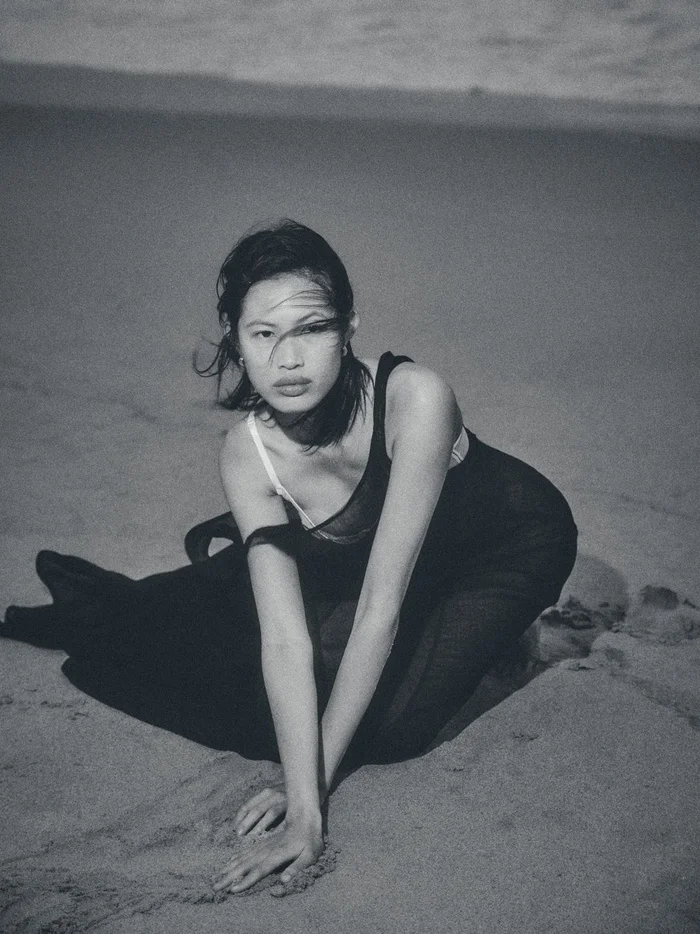 Monochrome portrait of an Asian woman in a black dress kneeling on a sandy beach with dark grainy film aesthetic