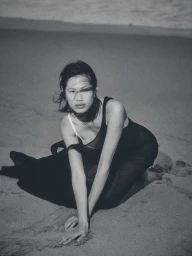 Monochrome portrait of an Asian woman in a black dress kneeling on a sandy beach with dark grainy film aesthetic