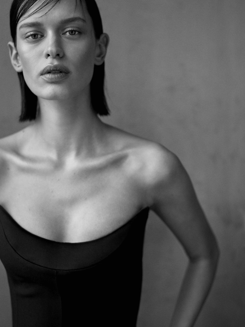 Monochromatic portrait of a woman in a black top with short hair, gazing directly at the camera against a blurred grey backdrop.