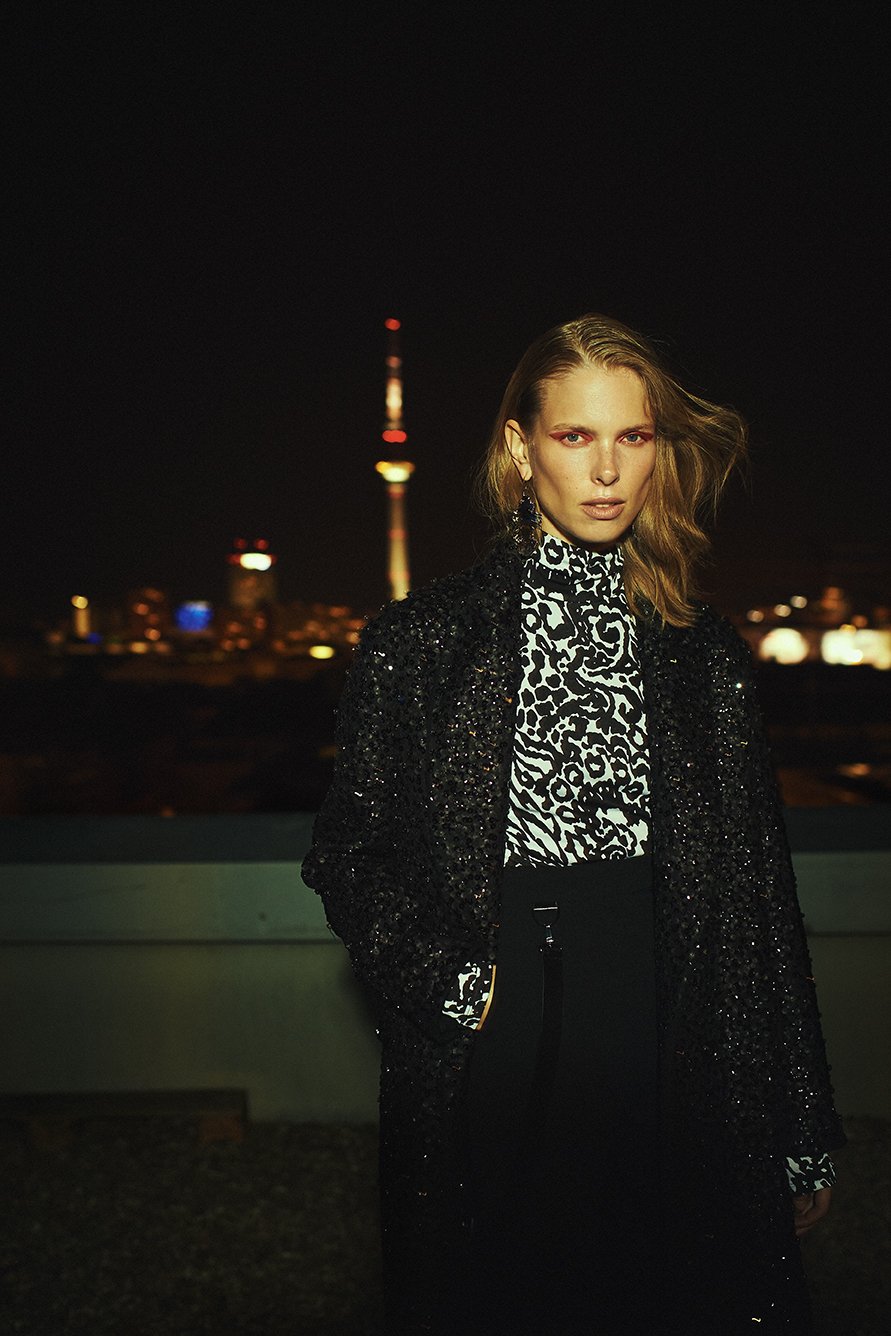A woman in a patterned shirt and black coat poses at night with city lights and a tall communications tower in the dark background.