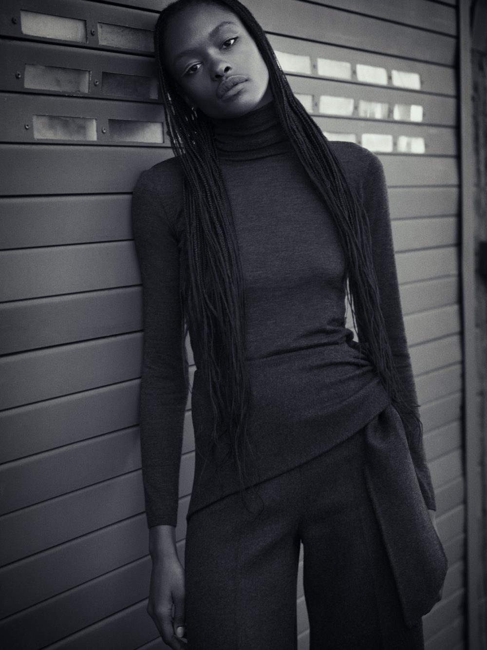 Monochrome portrait of a Black woman wearing a dark turtleneck and draped pants, leaning against a corrugated metal wall in a fashion shot.