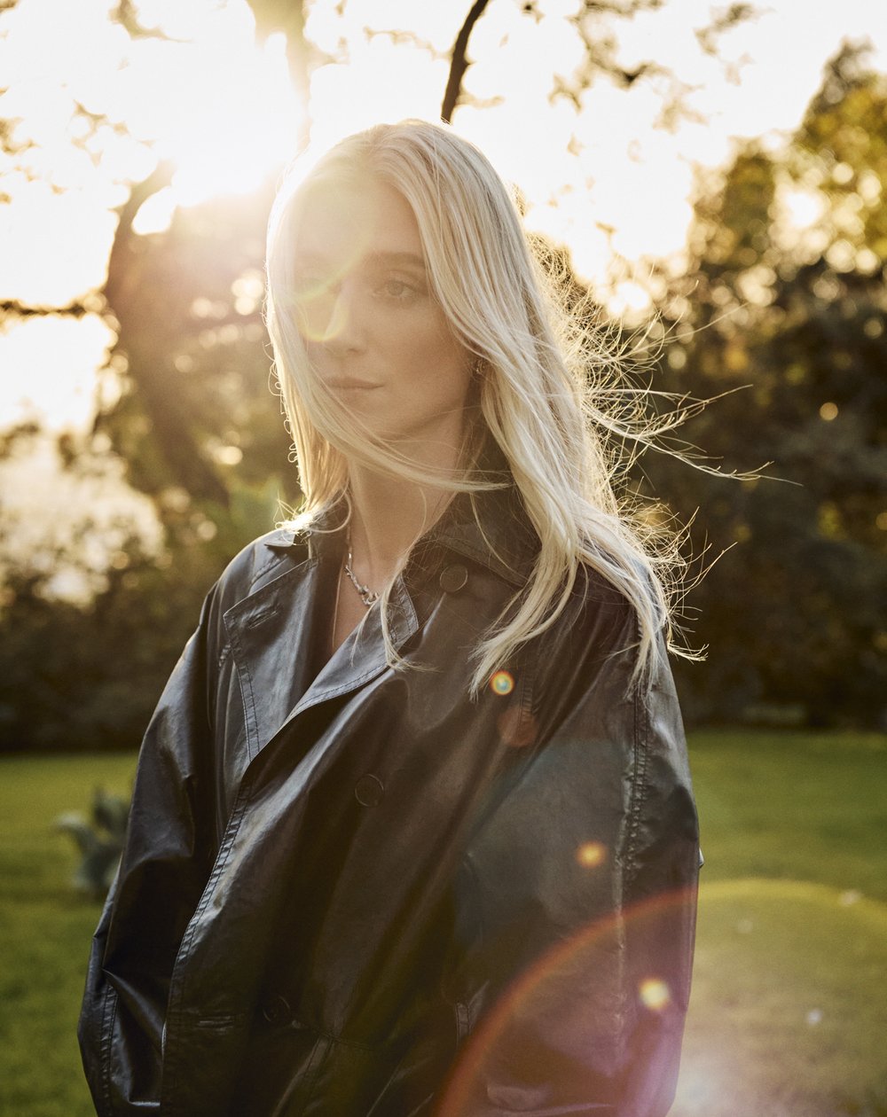 Portrait of a blonde woman in a black jacket standing outdoors in natural sunlight. Green foliage and dappled light fill the background.