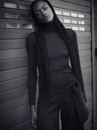 Black and white portrait of a woman with long braided hair wearing a dark turtleneck leaning against a shuttered door.