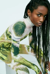 Black woman with long braided hair wearing a patterned green and white turtleneck top against a white background