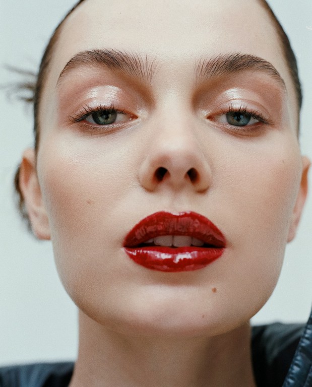 Close-up beauty shot featuring an adult female model with glossy red lipstick and dewy makeup against a plain white backdrop.