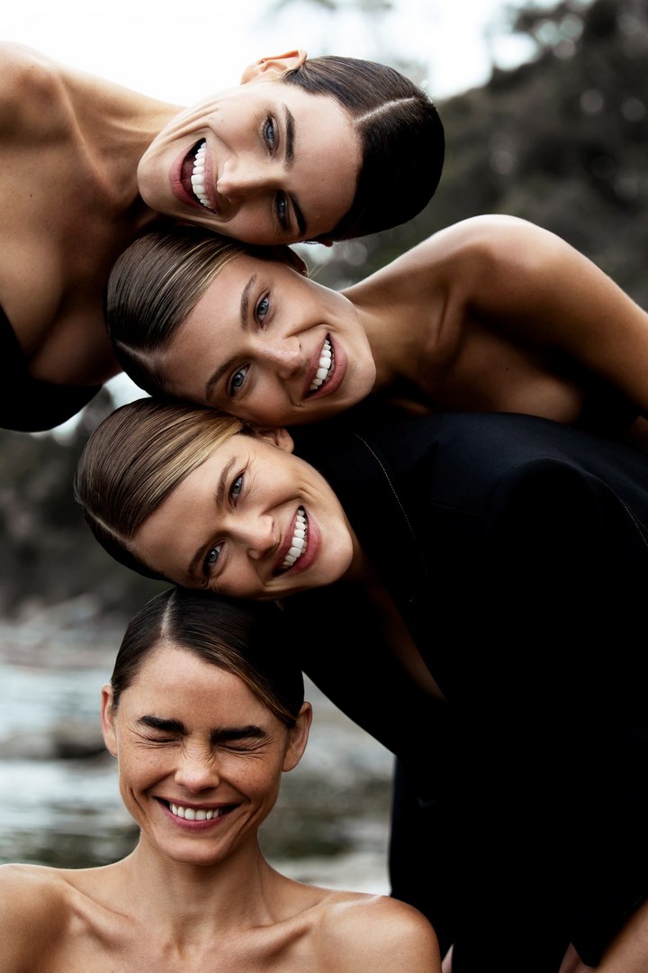 Close-up of four adult women smiling and stacked, wearing a mix of formal black jackets and bare shoulders, against an out-of-focus outdoor backdrop.