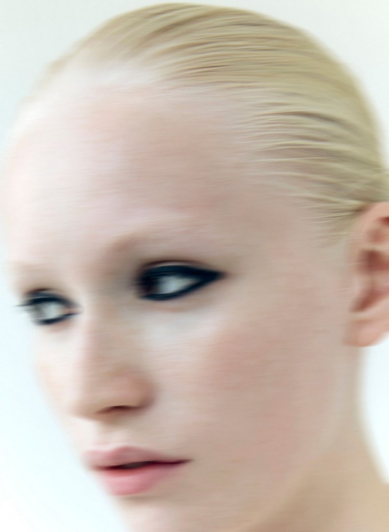 Close-up portrait of a woman with pale skin and striking dark eyeliner, soft focus, against a bright white background.