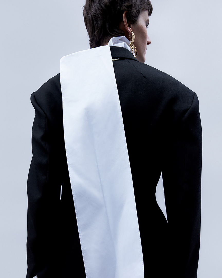 Model seen from behind wearing a black structured blazer with a long white fabric panel down the back against a white background