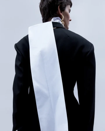 Model seen from behind wearing a black structured blazer with a long white fabric panel down the back against a white background