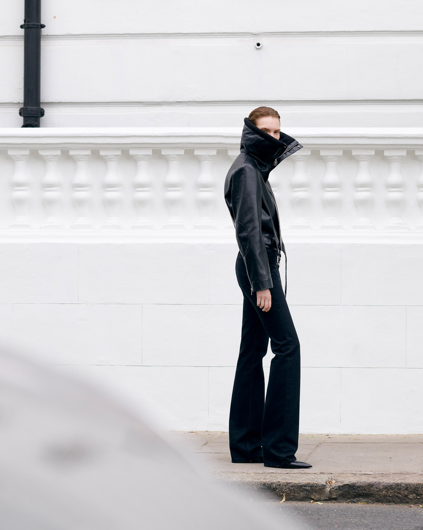 Fashionable woman in a black leather jacket and flared jeans stands confidently against a white wall, showcasing modern street style.