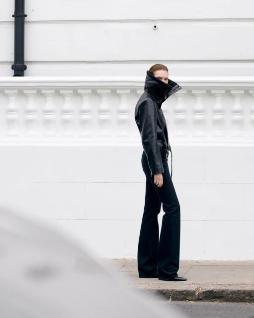 Fashionable woman in a black leather jacket and flared jeans stands confidently against a white wall, showcasing modern street style.
