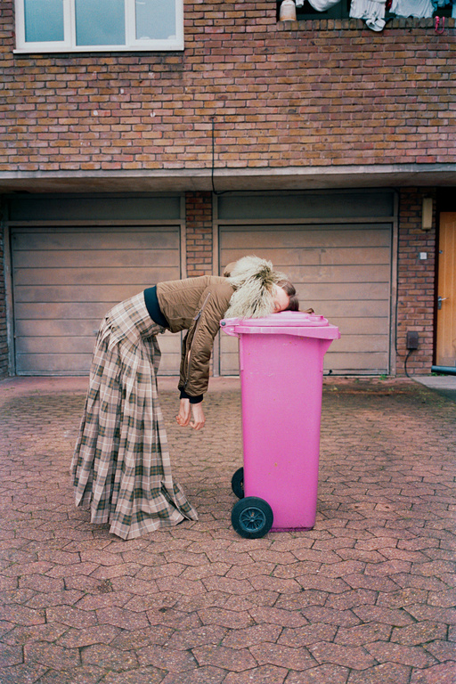 Woman in a plaid skirt leans on a pink trash can in an outdoor setting, showing a casual and unconventional street-style scene.