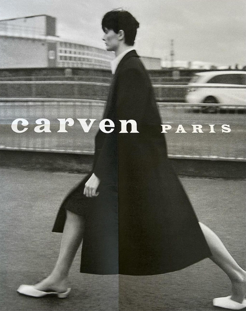 Monochrome outdoor fashion shot of a woman walking in a long coat and flat shoes against a backdrop of modern buildings and overcast skies.