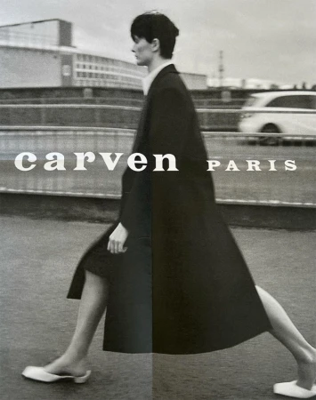 Monochrome outdoor fashion shot of a woman walking in a long coat and flat shoes against a backdrop of modern buildings and overcast skies.