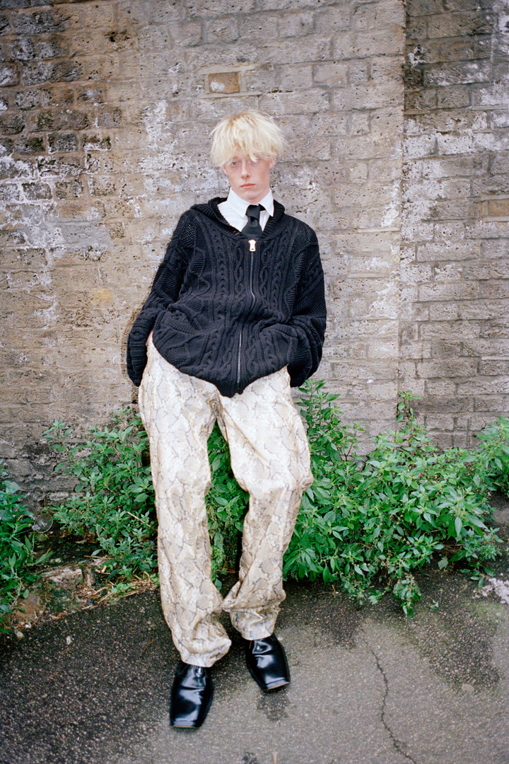 Young adult with blond hair stands against brick wall, wearing a black knit hooded cardigan, patterned pants, shiny black shoes outdoors.