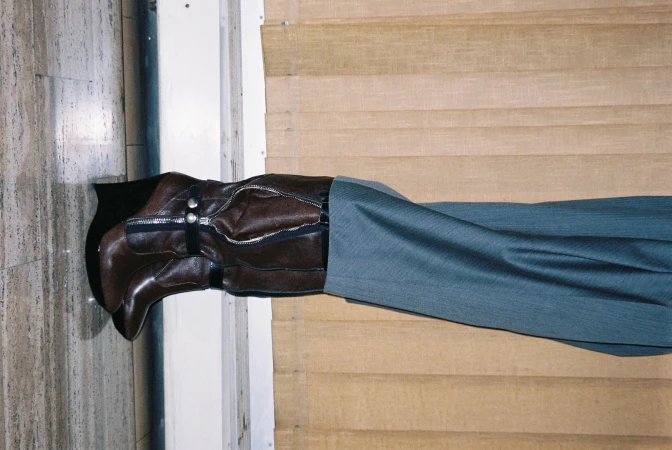 Brown leather knee-high boot with zipper and grey pants, presented against a light-brown wood paneled wall and marble flooring.