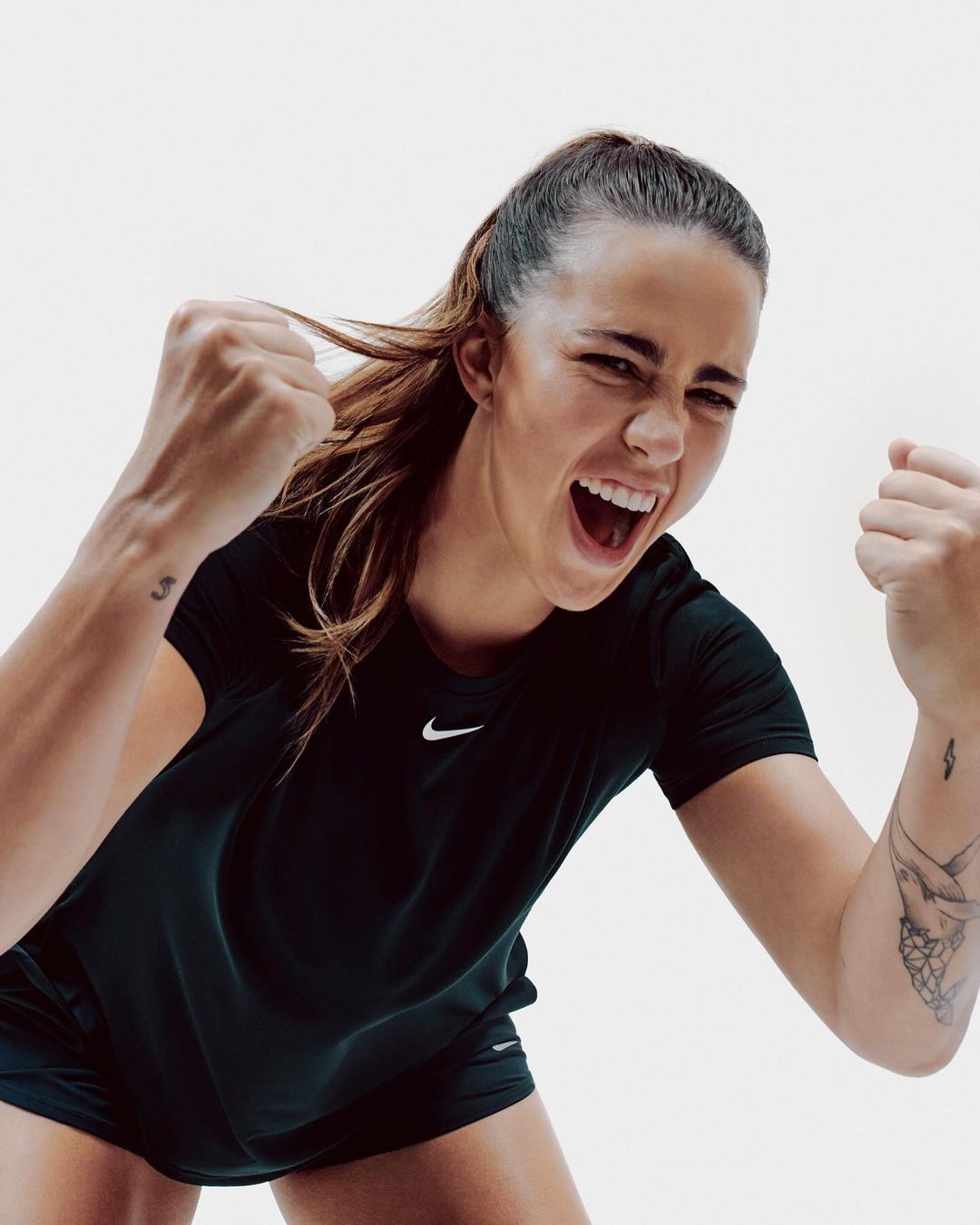 An athletic woman in workout clothing looks upwards and expresses energetic positivity with raised fists in a brightly lit indoor studio.