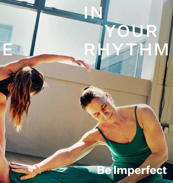 Two women in activewear practice yoga in a sunlit indoor space with large windows, promoting fitness and flexibility.