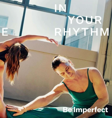 Two women in activewear practice yoga in a sunlit indoor space with large windows, promoting fitness and flexibility.
