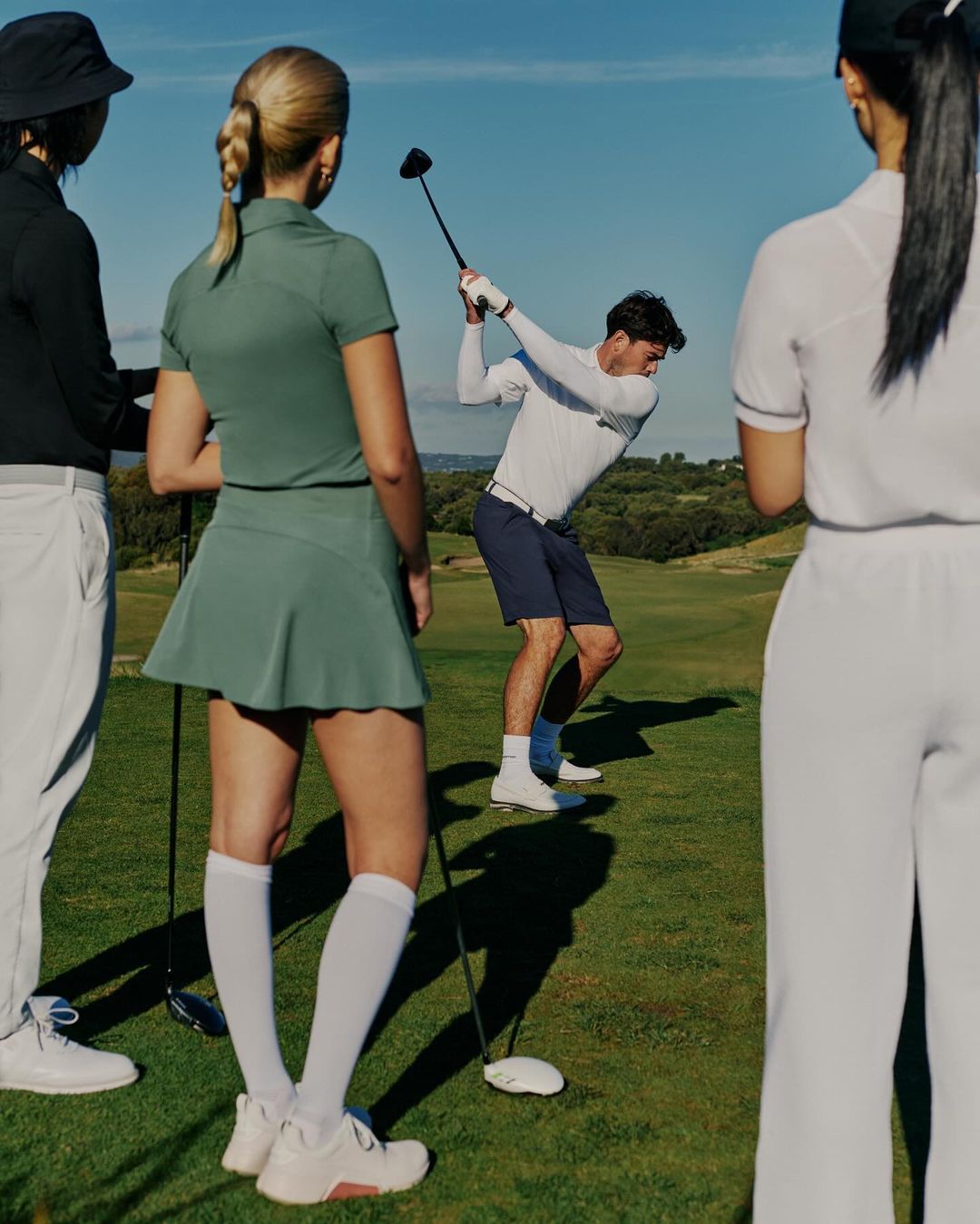 A group watches a man in white swing a golf club on a green golf course with a blue sky and trees in the background on a sunny day.