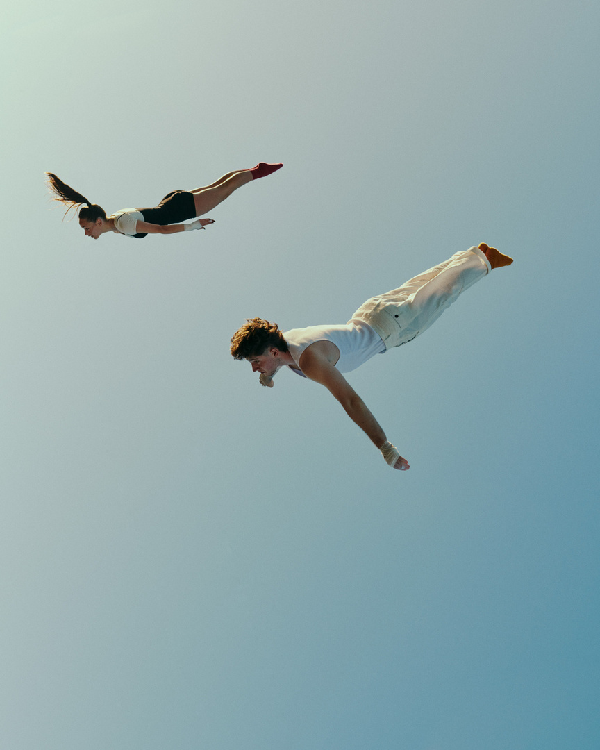 Woman and man in mid-air, wearing athletic clothing, limbs extended, against a clear sky, suggesting movement or flight in outdoor setting.