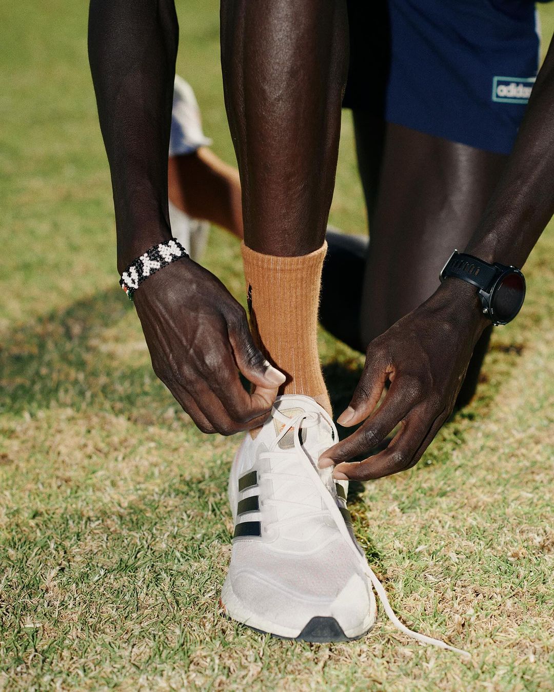 Runner tying the laces of an Adidas running shoe on a grass field, wearing a beaded bracelet and watch, with natural lighting.