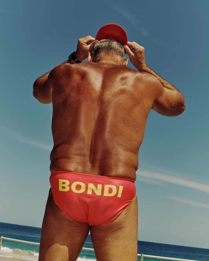 Back view of a mature man in a red swimsuit and red cap adjusting his cap, on a beach with blue sky, sunny day, Bondi written on suit.