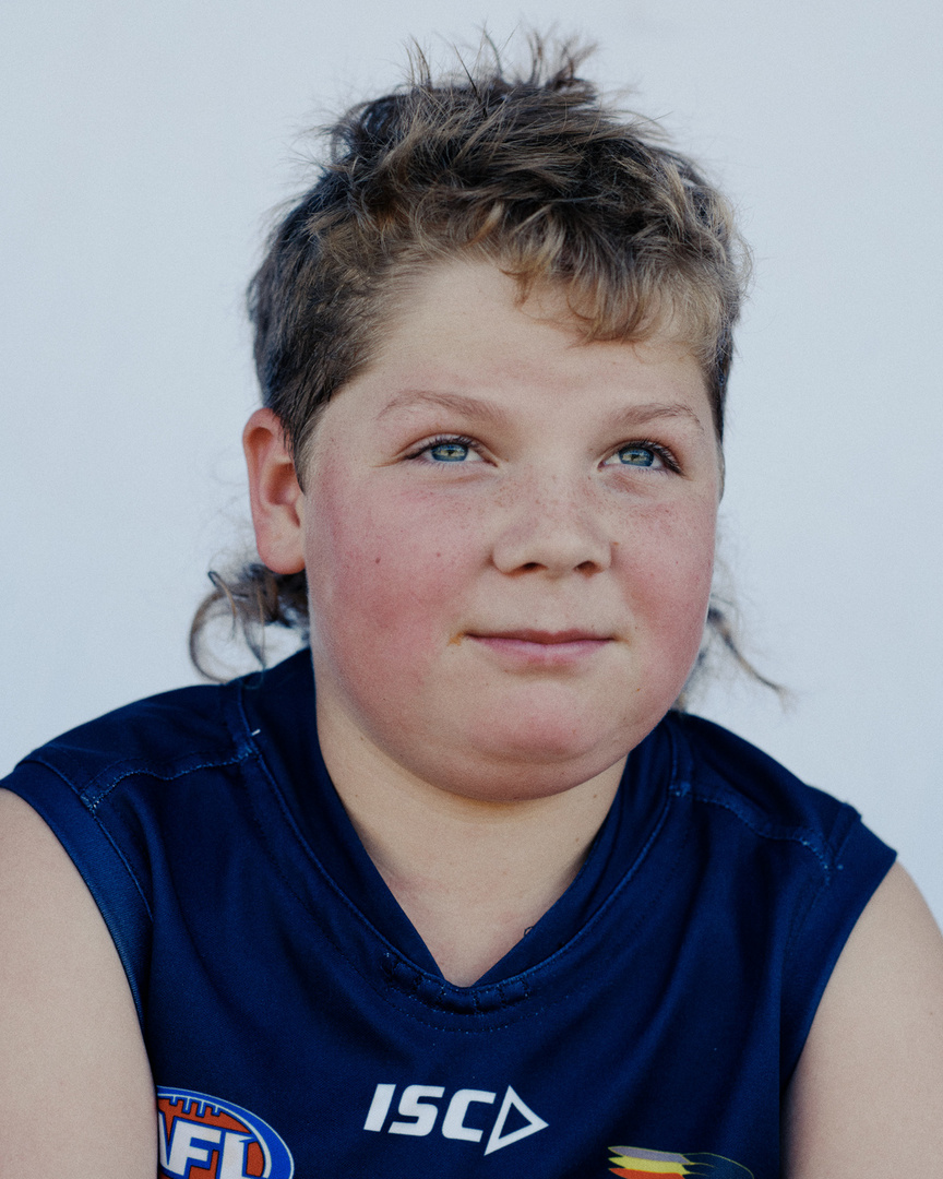 Portrait of a smiling boy with a mullet hairstyle wearing a sports jersey against a plain background, captured in natural light.