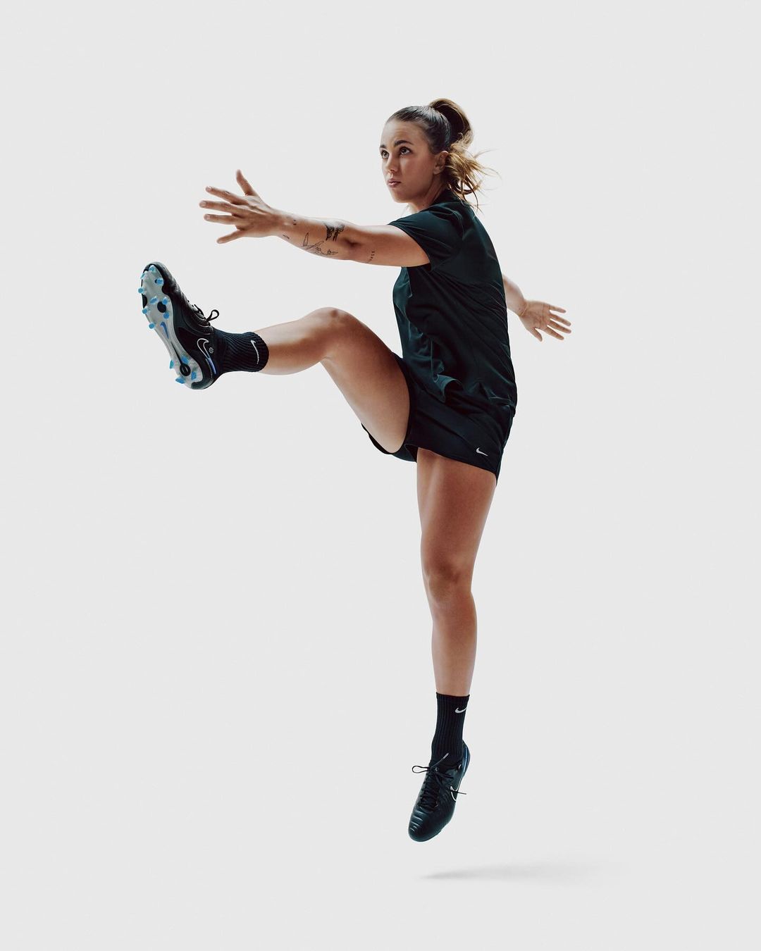 Athletic woman in motion wearing sports clothing and cleats, caught mid-air against a plain white backdrop in a dynamic studio shot.