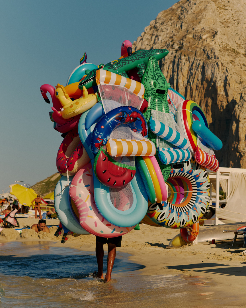 Person carries a stack of colorful inflatable pool floats on a beach with a mountain in the background on a sunny day.
