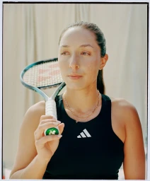 Portrait of a female tennis player in a black athletic top holding a racket over her shoulder against a neutral draped backdrop.