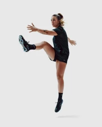Female athlete mid-air kicking in black sports kit and football boots against white backdrop by Chris Gurney