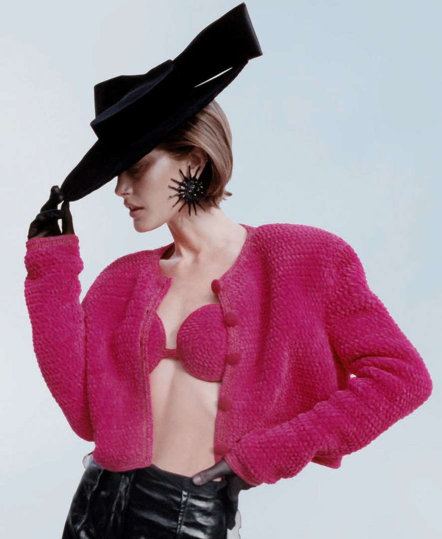 Woman in a hot pink jacket with a black hat and black gloves poses in front of a light blue background in a fashion studio shot.