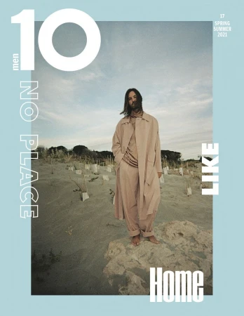 Magazine cover featuring a long-haired adult male in a tan outfit and coat, standing barefoot on a sandy, dune-like surface outdoors.