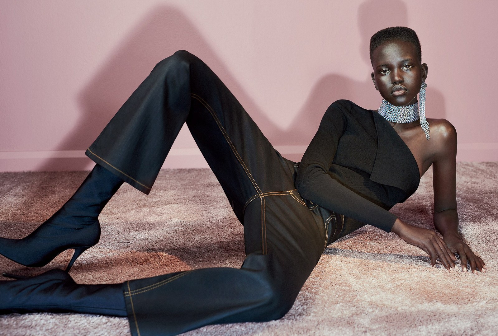Fashion shot of a woman in a black one-shoulder top, dark pants, and high-heeled boots, lying on a carpet in front of a pink wall.