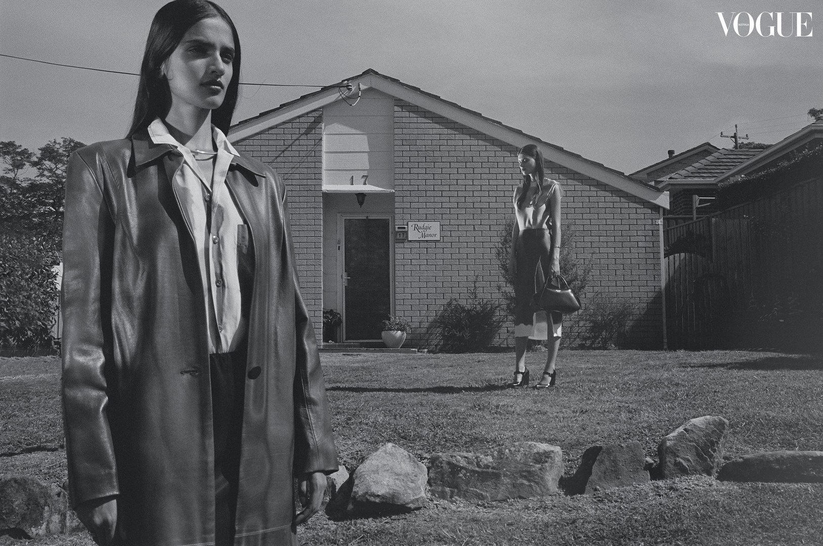 Black-and-white fashion shot with two women in stylish clothing outdoors in front of a brick house, Vogue logo in the upper right.