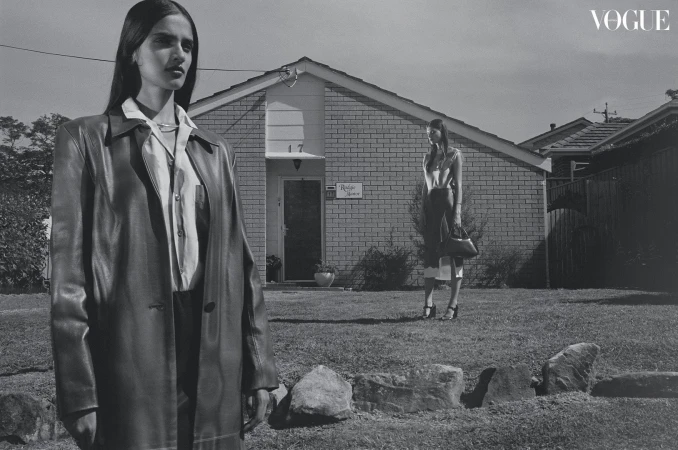 Black-and-white fashion shot with two women in stylish clothing outdoors in front of a brick house, Vogue logo in the upper right.