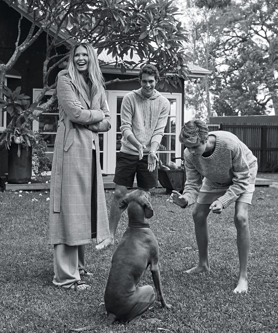 Black and white shot of woman in coat, two young men, and a dog in a grassy yard near a house, with trees visible in the background.
