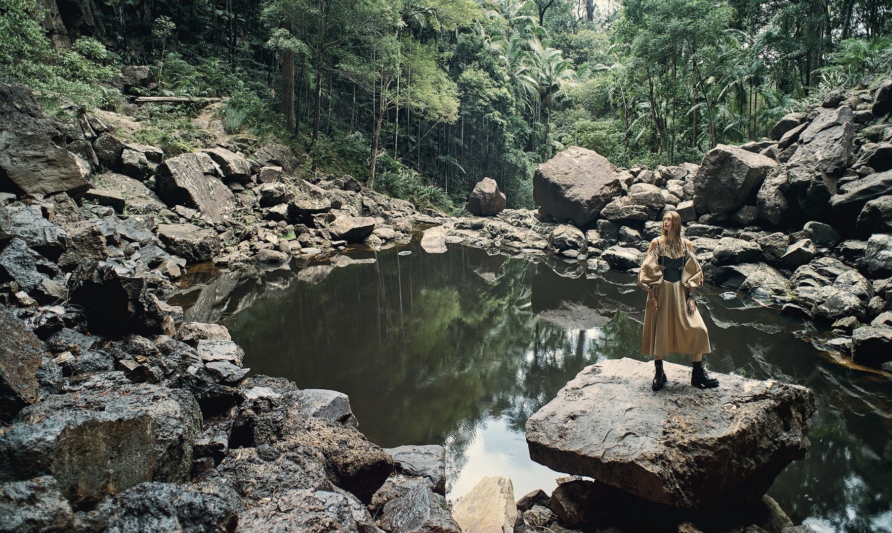 Woman in dress stands on rock in forest pool, surrounded by boulders and lush foliage. Trees are reflected in the water.