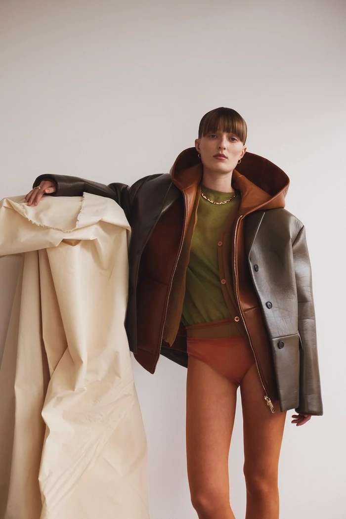 Model with bangs wearing an oversized olive leather jacket and green top resting arm on a large fabric roll against a white wall.