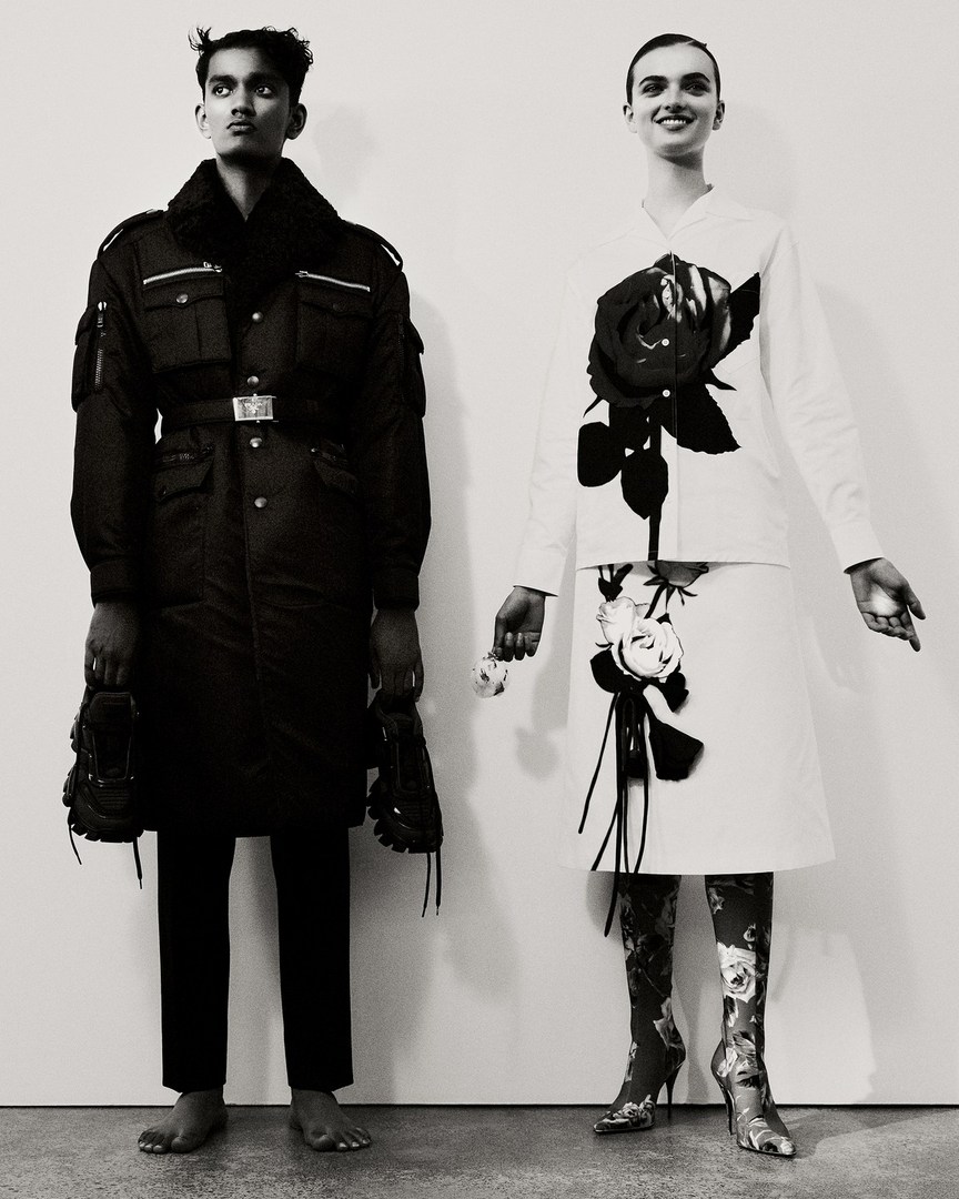 Two fashion models pose in a photo studio with black and white outfits, floral patterned boots, and bare feet against a white backdrop.
