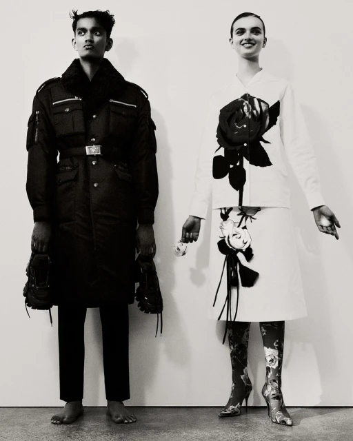 Two fashion models pose in a photo studio with black and white outfits, floral patterned boots, and bare feet against a white backdrop.