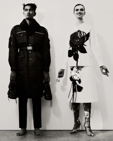 Two fashion models pose in a photo studio with black and white outfits, floral patterned boots, and bare feet against a white backdrop.